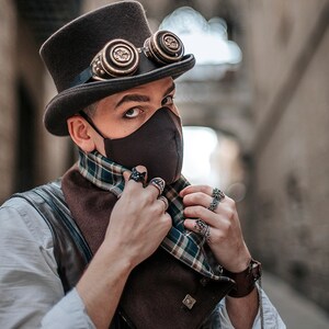 Puede incluir: Una persona con una m&aacute;scara facial negra, un sombrero de copa marr&oacute;n con gafas, un chaleco de cuero marr&oacute;n, una camisa blanca y una bufanda a cuadros. La persona est&aacute; ajustando la bufanda con sus manos, que est&aacute;n adornadas con varios anillos.