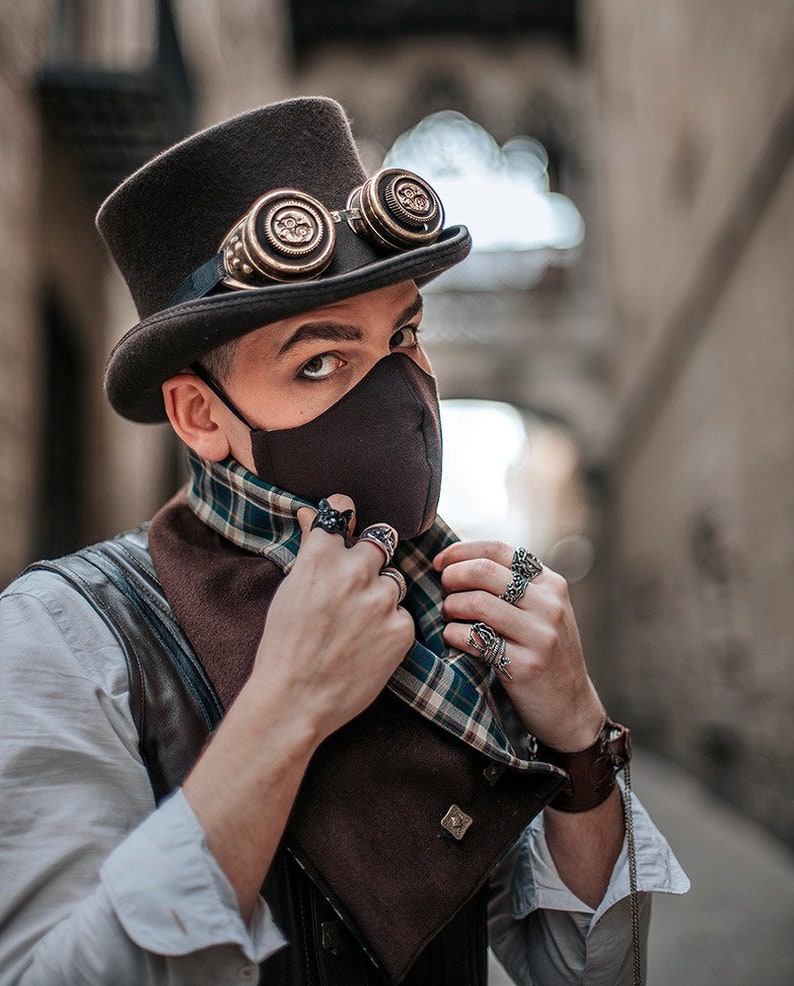 Puede incluir: Una persona con una m&aacute;scara facial negra, un sombrero de copa marr&oacute;n con gafas, un chaleco de cuero marr&oacute;n, una camisa blanca y una bufanda a cuadros. La persona est&aacute; ajustando la bufanda con sus manos, que est&aacute;n adornadas con varios anillos.