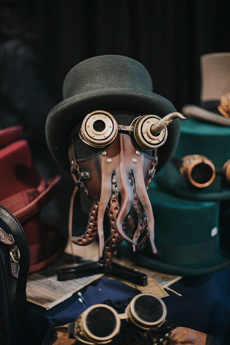 May include: A steampunk-style mask with a top hat, goggles, and an octopus-like design. The mask is made of brown leather and has a dark green top hat. The goggles are made of brass and have round lenses. The octopus tentacles are made of brown leather and have a textured surface.