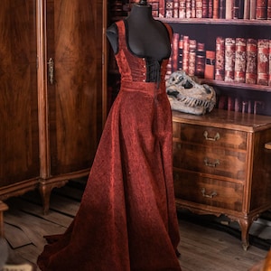 Op de afbeelding: Een rode fluwelen jurk met een zwart kanten korset en een lange rok. De jurk is op een mannequin in een kamer met een houten kast en een boekenkast vol boeken.