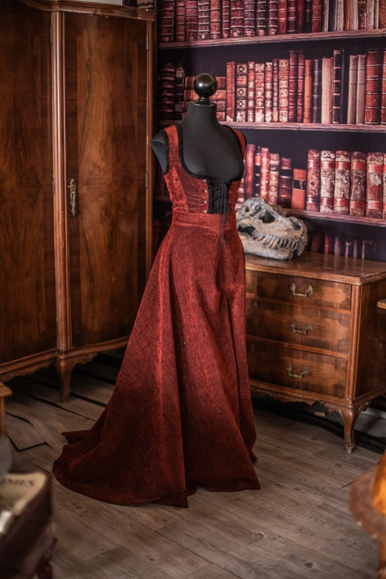 Op de afbeelding: Een rode fluwelen jurk met een zwart kanten korset en een lange rok. De jurk is op een mannequin in een kamer met een houten kast en een boekenkast vol boeken.