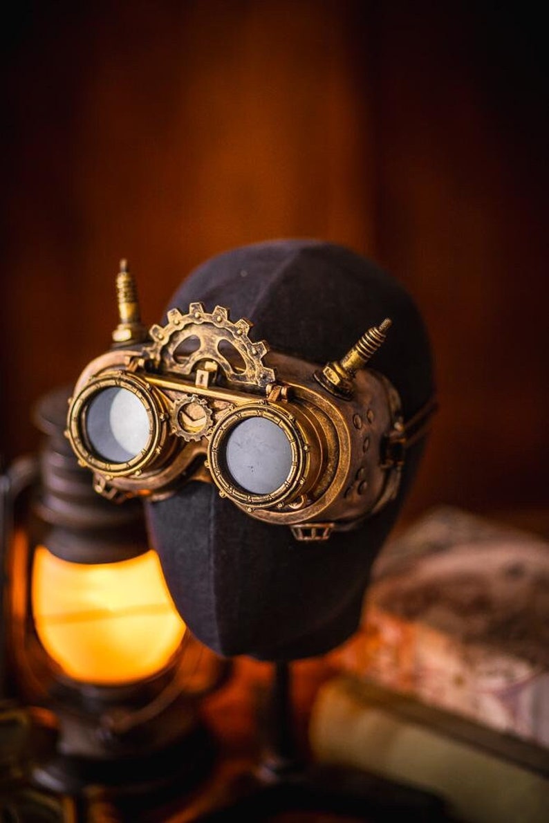 May include: Brass-colored goggles featuring two circular lenses, a central gear mechanism, and two small vertical spikes on the top frame. The goggles are mounted on a dark, featureless head-shaped display form with a metallic, weathered finish.