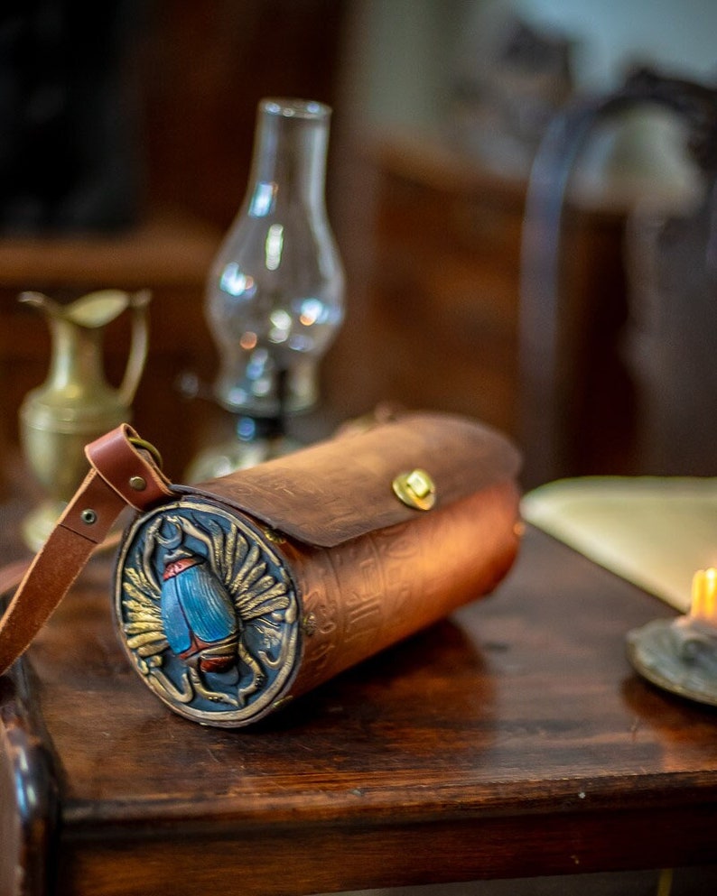 May include: A brown leather cylindrical bag with a strap. The bag has a circular front panel with a gold-colored scarab beetle design with blue wings and a red body.