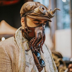 May include: A person wearing a brown leather cap with goggles and a brown leather octopus mask. The mask has a long, curled tentacle extending from the side. The person is wearing a brown tweed jacket over a white shirt with a map print.