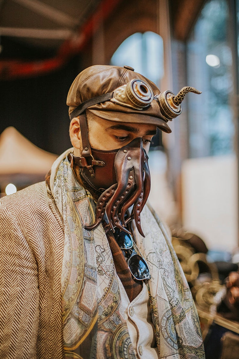 May include: A person wearing a brown leather cap with goggles and a brown leather octopus mask. The mask has a long, curled tentacle extending from the side. The person is wearing a brown tweed jacket over a white shirt with a map print.