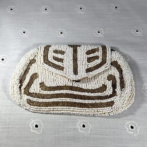 May include: A vintage beaded clutch purse in ivory and brown. The purse features a geometric design on the front flap and body. The clutch is rectangular with a curved top and a clasp closure. The background is a white fabric with a floral pattern.