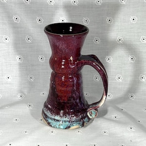 May include: A handmade ceramic mug with a deep burgundy glaze. The mug features a flared top, a bulbous midsection, and a turquoise and white base. The handle is curved and the interior is a dark, glossy color. This unique mug is perfect for coffee or tea.