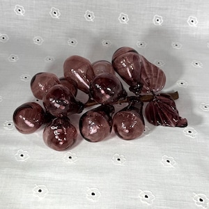 May include: A cluster of purple glass grapes with a brown stem and leaves. The grapes have a glossy finish and vary in size. The decorative piece is set against a white background with a floral pattern.