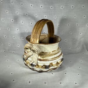 May include: A handcrafted ceramic basket with a curved handle and a decorative bow on the side. The basket features a speckled, off-white glaze with brown and tan stripes and accents. The rim and handle are a darker brown.