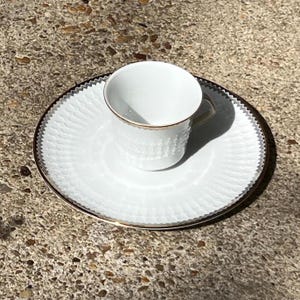 May include: A white teacup and saucer set. The cup and saucer have a textured pattern and gold trim. The saucer has a scalloped edge. The set is on a stone surface.