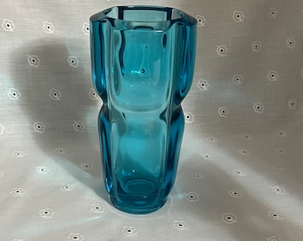May include: A turquoise glass vase with a unique, curved design. The vase has a rectangular shape with rounded sides and a wide opening. The glass is transparent, allowing light to pass through. The vase is a decorative item.