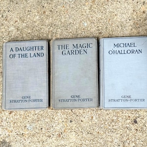 May include: Three vintage hardcover books with light gray fabric covers. The titles are A Daughter of the Land, The Magic Garden, and Michael O'Halloran, all authored by Gene Stratton-Porter, with black text printed on the front covers.