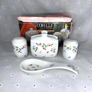 May include: A Corelle Coordinates table set featuring a white ceramic napkin holder, salt and pepper shakers, and a spoon rest, all decorated with a pattern of small red, yellow, and blue flowers with green leaves, presented in its original box.