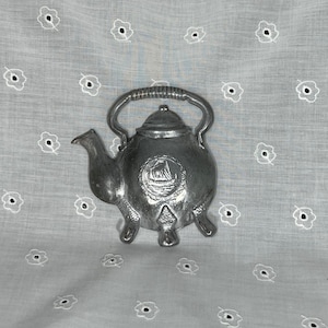 May include: A silver-colored teapot-shaped decorative item. The teapot has a handle, a spout, and three feet. A ship emblem is embossed on the front. The background is a white fabric with small, embroidered flower patterns.