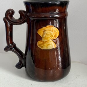 May include: A brown ceramic mug with a raised design of a person's head wearing a hat. The mug has a dark brown glaze and a handle with a decorative design.