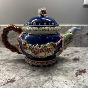 Puede incluir: Una tetera de cerámica azul y marrón con un diseño floral y un asa retorcida.
