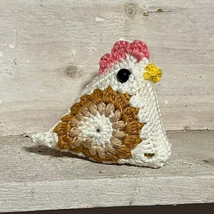 May include: A crocheted chicken toy with a white body, brown and beige patterned wings, a pink comb, a yellow beak, and black eyes.