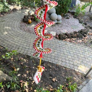 May include: A handmade, spiral-shaped crochet ornament. The ornament features a red central cord with white, green, and gold crochet detailing. A small, matching crochet piece hangs at the bottom. The ornament is suspended outdoors.