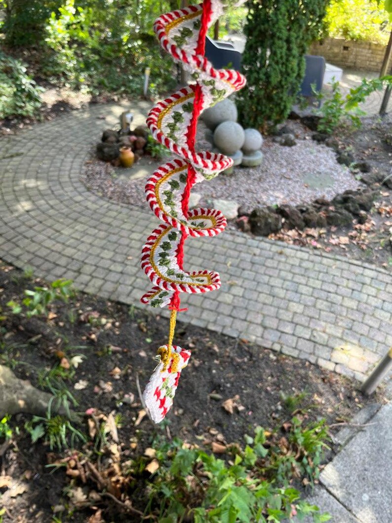 May include: A handmade, spiral-shaped crochet ornament. The ornament features a red central cord with white, green, and gold crochet detailing. A small, matching crochet piece hangs at the bottom. The ornament is suspended outdoors.