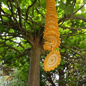 Peut inclure: Ornement décoratif en crochet jaune en forme de spirale suspendu à une branche d'arbre. L'ornement présente un motif circulaire avec un motif ajouré détaillé. L'arrière-plan présente un arbre avec des feuilles vertes et de la lumière du soleil.