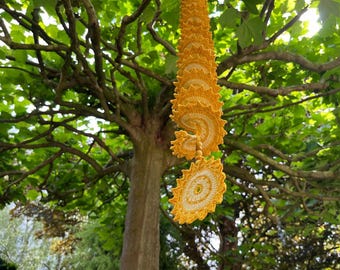 May include: A hanging spiral decoration crafted from yellow and cream-colored crochet yarn. The piece features a series of interconnected, circular, sun-like motifs with scalloped edges that cascade downward in a continuous, twisting vertical line.