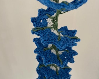 Peut inclure: Une guirlande crocheté de fleurs bleues, blanches et vertes. La guirlande est faite de fil et est enfilée avec un fil vert.
