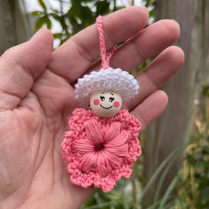 Puede incluir: Una flor de crochet rosa con un sombrero de crochet blanco y una cara de madera con ojos pintados y una sonrisa.