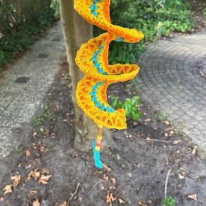 Könnte beinhalten: Eine handgefertigte, spiralförmige Häkeldekoration in leuchtendem Gelb und Orange mit türkisfarbenen Akzenten. Die Spirale hängt an einem Baum und hat eine türkisfarbene Quaste mit bunten Perlen am unteren Ende.