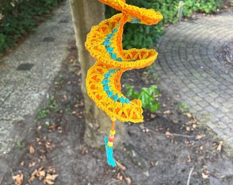 Puede incluir: Una decoración de ganchillo en forma de espiral, hecha a mano, en amarillo brillante y naranja, con detalles turquesa. La espiral cuelga de un árbol y tiene una borla turquesa con cuentas de colores en la parte inferior.