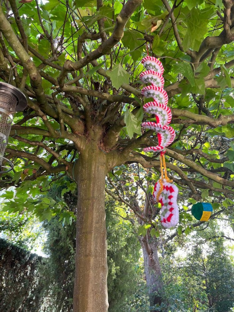 May include: A tree with green leaves and a brown trunk. Hanging from the branches are two crocheted spiral ornaments with red and white stripes. A yellow string and a green, blue, and yellow geometric ornament are also hanging from the tree.