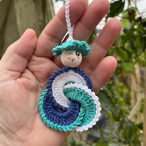 Op de afbeelding: Handgemaakt gehaakt ornament met een glimlachend houten kralen gezicht en een turquoise hoed. Het ornament heeft een blauw, wit en turquoise spiraalontwerp. Een wit touwtje is bevestigd om op te hangen. Een grillig item.