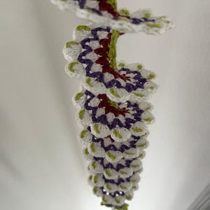 May include: A handmade crochet spiral decoration. The spiral features white, purple, red, and green crochet elements. The decorative piece is suspended from the ceiling, showcasing its intricate design and color palette.