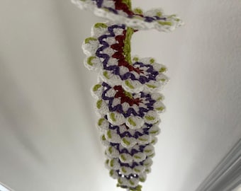 Könnte beinhalten: Eine handgefertigte Häkelspirale. Die Spirale besteht aus weißen, lila, roten und grünen Häkelelementen. Das dekorative Stück hängt von der Decke und zeigt sein kompliziertes Design und seine Farbpalette.