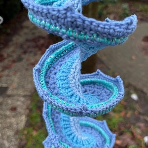 May include: A spiral-shaped, handmade crochet piece in shades of blue and teal. The crochet work features a textured pattern with a scalloped edge. The piece is suspended, showcasing its three-dimensional form and intricate details.