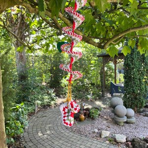 May include: A handmade crochet spiral mobile with red and white stripes, suspended from a tree branch. The mobile features a spiral design and a red hanging cord. The bottom of the mobile has a crochet design with red and white colors.