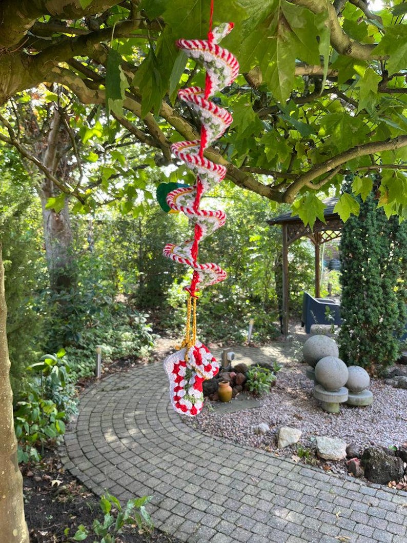 May include: A handmade crochet spiral mobile with red and white stripes, suspended from a tree branch. The mobile features a spiral design and a red hanging cord. The bottom of the mobile has a crochet design with red and white colors.