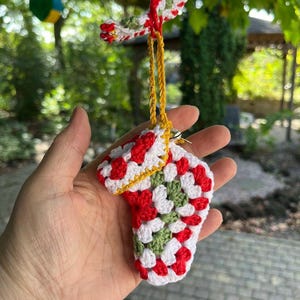 May include: A handmade crochet Christmas stocking ornament in red, white, and green. The small stocking is suspended by a gold cord. The ornament is made of granny squares and has a white border.