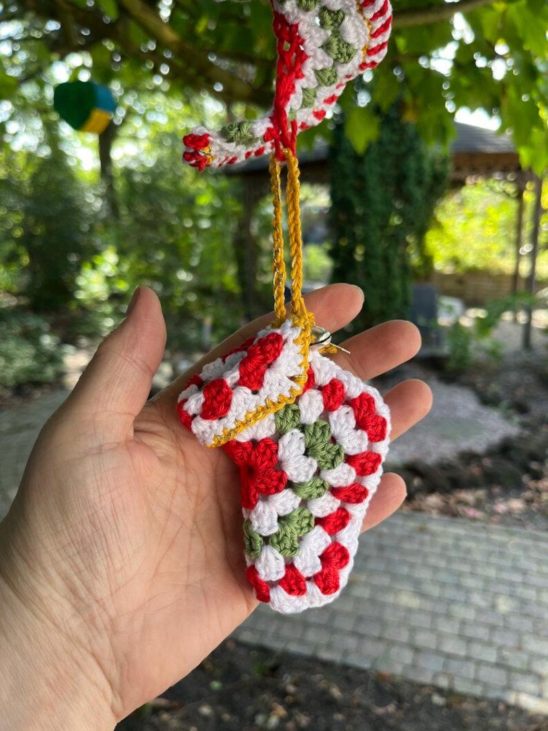 May include: A handmade crochet Christmas stocking ornament in red, white, and green. The small stocking is suspended by a gold cord. The ornament is made of granny squares and has a white border.