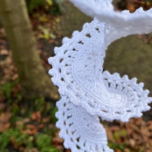 Könnte beinhalten: Nahaufnahme eines weißen, gehäkelten Schals mit einem gewellten Rand. Der Schal ist aus weichem, strukturiertem Garn gefertigt und in Spiralform drapiert. Der Hintergrund ist verschwommen, mit einem Baumstamm und fallenden Blättern.