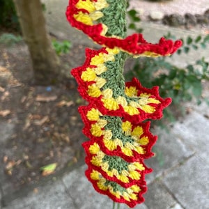 May include: A spiral-shaped crochet hanging decoration featuring a central green column with attached ruffled layers in yellow and red. The structure twists downward, showcasing a repeating pattern of crocheted circles with yellow centers and red edges.