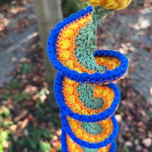 May include: A colorful, crocheted spiral decoration. The spiral features a vibrant combination of blue, orange, yellow, and green yarn. The design is reminiscent of a stylized flower or abstract art, with a textured, handmade appearance.
