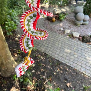May include: A handmade, spiral-shaped crochet ornament with red, white, green, and gold accents. The ornament is suspended outdoors, showcasing a festive design. The crochet work features a repeating pattern, creating a visually appealing, decorative piece.