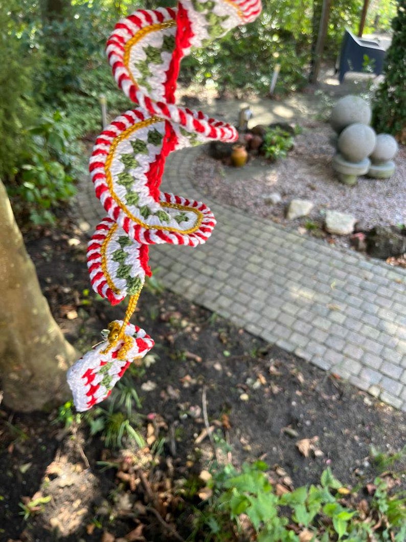 May include: A handmade, spiral-shaped crochet ornament with red, white, green, and gold accents. The ornament is suspended outdoors, showcasing a festive design. The crochet work features a repeating pattern, creating a visually appealing, decorative piece.