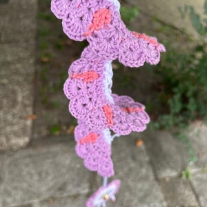 May include: A spiral-shaped crochet hanging decoration featuring a white central spine with lavender scalloped edges and small coral-colored accents. The structure twists downward, ending in a small, circular lavender and white crocheted flower charm.