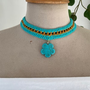 Op de afbeelding: Een turquoise en gouden gehäkelde choker ketting met een turquoise bloemenhanger.