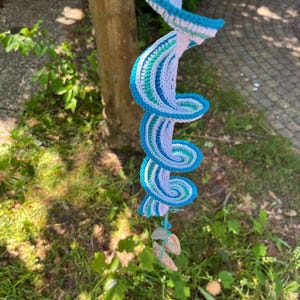 Op de afbeelding: Een handgemaakte gehaakte spiraal in tinten blauw, wit en groen, hangend aan een boom. Het spiraalontwerp heeft een reeks geneste curven, met een franje van kleine, natuurlijk gekleurde stenen aan de onderkant. Het stuk is geplaatst tegen een achtergrond van groen gras en een boomstam.