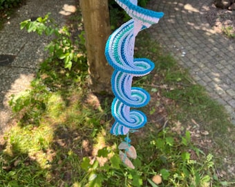 May include: A handmade crochet spiral in shades of blue, white, and green, hanging from a tree. The spiral design features a series of nested curves, with a fringe of small, natural-colored stones at the bottom. The piece is set against a backdrop of green grass and a tree trunk.