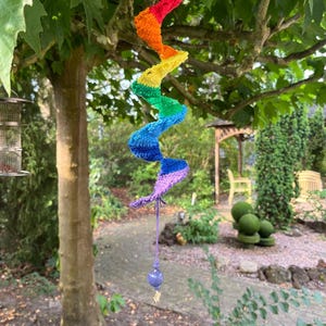 Puede incluir: Una decoración colgante de ganchillo en forma de espiral con un degradado de colores arcoíris, desde el rojo en la parte superior hasta el morado en la inferior. Un cordón morado cuelga de la base con una cuenta morada y un dije metálico.