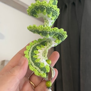 May include: A green and white crocheted spiral with a green and white beaded tassel. The spiral is made of multiple layers of crocheted fabric.