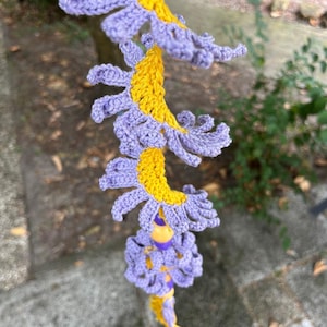 May include: A vertical hanging decoration featuring a series of crocheted flower-like shapes in bright yellow and light purple yarn. The structure is spiraled, with purple petals extending from a yellow center, accented by small purple and yellow beads.